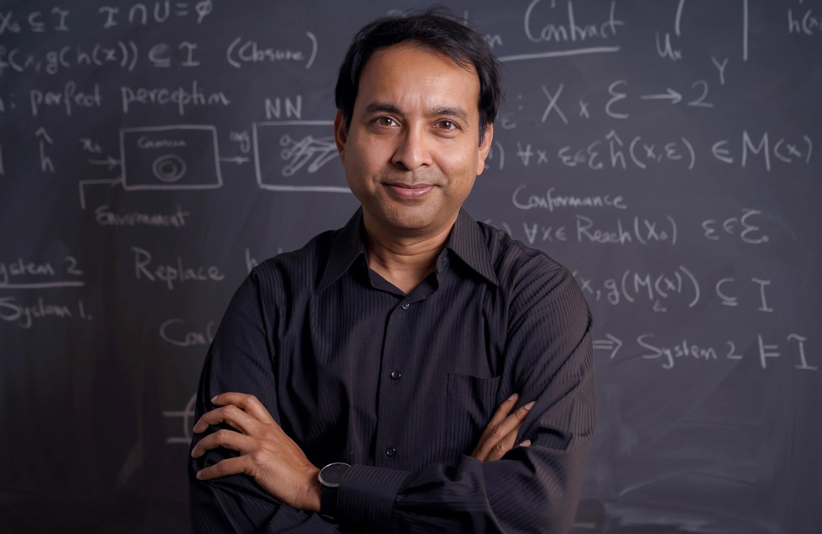 Person wearing a dark grey button down shirt and crossing their arms while posing in front of a dark grey chalkboard...